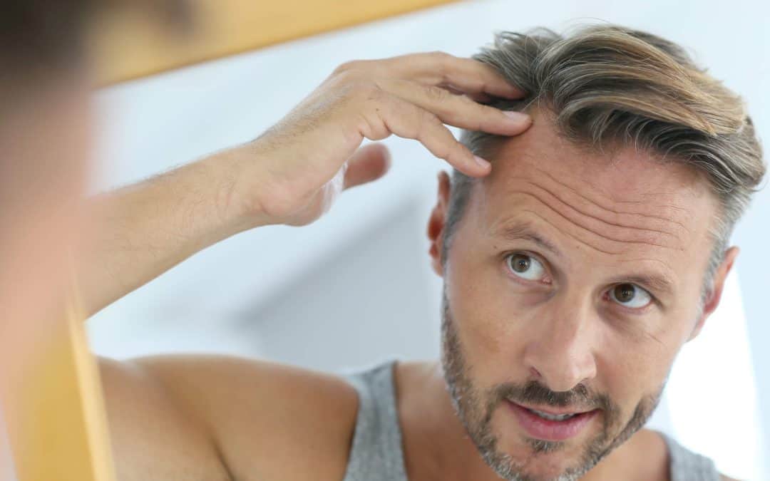 A man with short hair and a beard examining his hairline in a mirror while wearing a gray tank top.